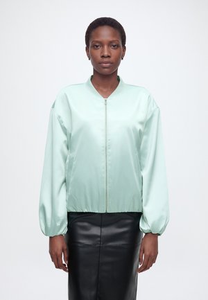 Femme aux cheveux courts portant une veste à fermeture éclair vert clair et une jupe en cuir noire, debout devant un fond blanc uni.