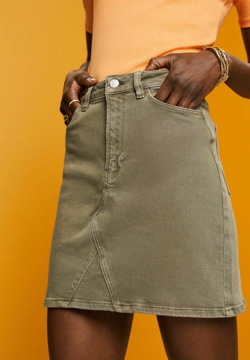 Khaki Denim-Minirock mit vorderem Knopfverschluss, Taschen und Seitennähten. Vor einem gelben Hintergrund.