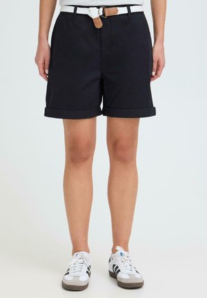 Person iført sorte opsmøgede shorts med hvid flettet bælte og hvide sneakers med sorte striber, stående mod en ensfarvet baggrund.