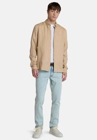 Camicia beige a maniche lunghe con colletto a bottoni, indossata sopra una maglietta bianca. Jeans blu chiaro e sneaker color crema completano l'outfit.