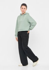 Giovane donna in piedi con una mano in tasca, che indossa un hoodie verde salvia, jeans neri a gamba larga e sneakers bianche su sfondo bianco.