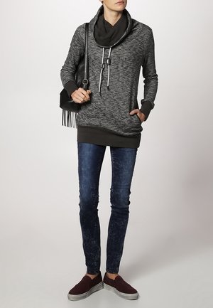 Haut à capuche marbré noir et gris avec un col en cowl et des cordons de serrage, associé à un jean skinny bleu foncé et des chaussures à enfiler bordeaux. Sac noir avec des franges.