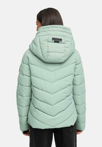 Woman wearing a light green quilted puffer jacket with a hood and black pants, shown from the back against a plain background.