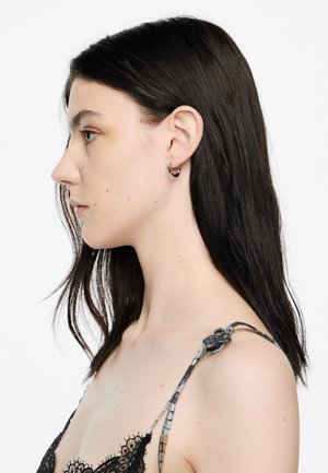 Pendientes de aro negros en una modelo con cabello lacio oscuro. La parte superior presenta un delicado diseño de encaje y tirantes estampados.