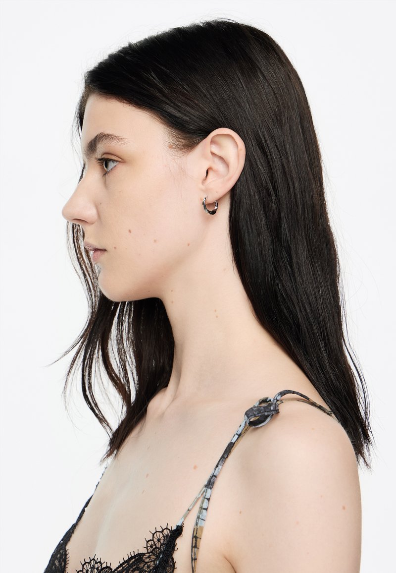 Pendientes de aro negros en una modelo con cabello lacio oscuro. La parte superior presenta un delicado diseño de encaje y tirantes estampados.