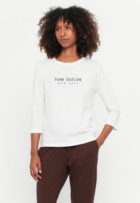 Witte lange mouw shirt met een ronde halslijn en marineblauwe tekst "TOM TAILOR EST. 1962." Gecombineerd met bruine broek, wat casual kleding toont.