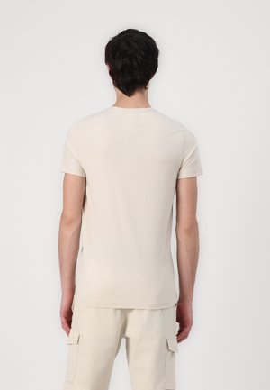 Homme aux cheveux bruns bouclés portant un t-shirt beige uni à manches courtes et un pantalon cargo beige, face à un mur blanc.