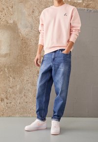 Sudadera de color rosa claro con un pequeño logo, combinada con jeans de mezclilla azul de corte holgado y zapatillas blancas con acentos rosas.