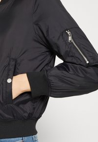 Blouson aviateur noir en tissu lisse et brillant, avec poignets côtelés, poches latérales et poche zippée sur la manche.