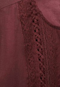Blusa burdeos con detalles de encaje a lo largo de un lado, que presenta un patrón intrincado y una textura que aporta profundidad.