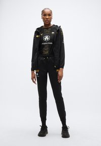 Tenue de sport noire comprenant une veste zippée à capuche, un t-shirt graphique et un pantalon de survêtement fuselé, tous ornés de logos dorés.