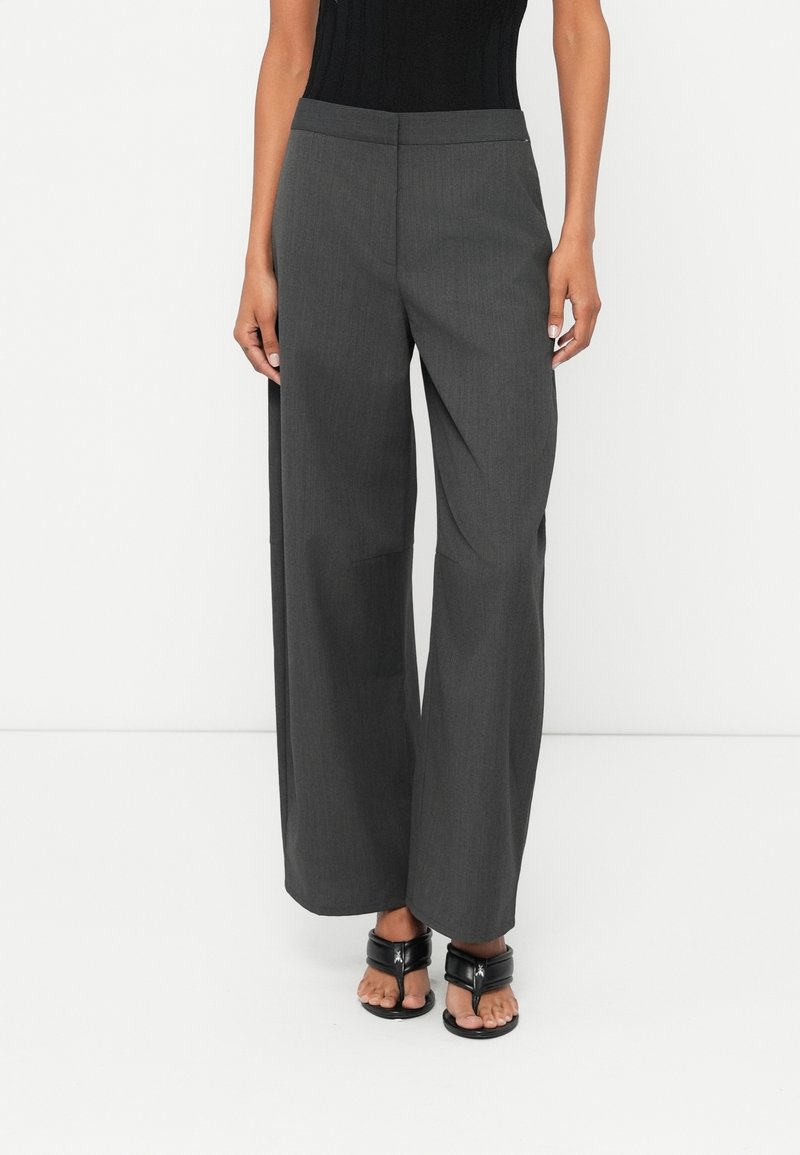 Femme portant un pantalon large gris foncé et des sandales à talons noires à bout ouvert sur fond blanc uni.