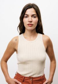 Débardeur beige à côtes avec un col haut et un design sans manches, associé à un pantalon couleur rouille. Les détails incluent une texturation subtile.