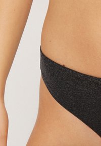 Černé bikinové kalhotky vyrobené z lesklého materiálu, s vysokým střihem, širokým pasem a minimálním pokrytím po stranách.