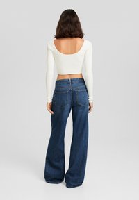 Top corto a maniche lunghe bianco a coste con una ampia scollatura a forma di ciotola sul retro, abbinato a jeans scuri a vita alta, larghi, con tasche posteriori.