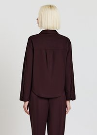 Blouse bordeaux avec un col, coupe décontractée et manches longues. Présente un pli au dos et des poignets à boutons pour un ajustement modulable.