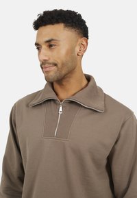 Pullover marrone con zip, colletto a coste e pannello testurizzato. Presenta una zip argentata e un tessuto morbido e liscio.