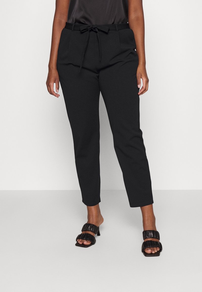 Pantalon noir fuselé pour femme avec taille à cordon, associé à des sandales à talons noires avec lanière froncée et orteils ouverts.