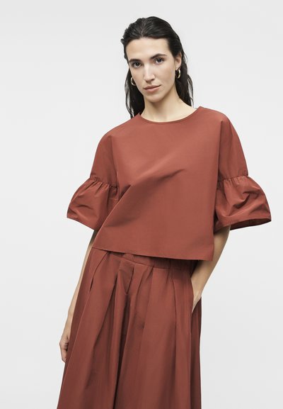 Γυναίκα με σκούρα μαλλιά που φοράει τοπ σε χρώμα σκουριάς με φουσκωτά μανίκια και ταιριαστή πλισέ φούστα με ψηλή μέση, στέκεται μπροστά σε ανοιχτό γκρι φόντο.