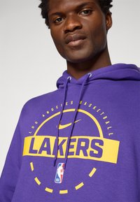Purple hoodie featuring "LAKERS" in yellow, with a circular design and Nike logo. Soft fabric with drawstrings and front graphic detail.
