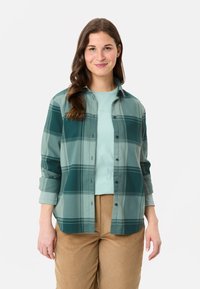 Grünes karriertes Hemd mit langen Ärmeln, über einem hellblauen T-Shirt getragen, kombiniert mit beige Kordhosen.