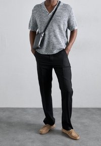 Osoba ubrana w szary, dziergany top z krótkim rękawem, czarne spodnie, beżowe zamszowe mokasyny i trzymająca małą czarną torbę przez ramię, stojąca na szarej podłodze.