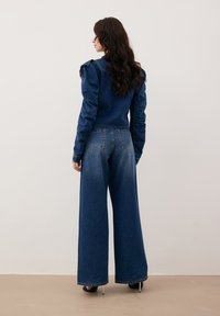 Kurtka dżinsowa z detalami na ramionach, w połączeniu z szerokimi niebieskimi dżinsami. Obie rzeczy mają klasyczne ciemne zabarwienie i strukturalny fason.