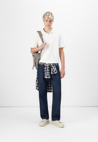 Young man with short blonde hair wearing a white polo, dark jeans, beige sneakers, plaid shirt tied around waist, and gray shoulder bag.