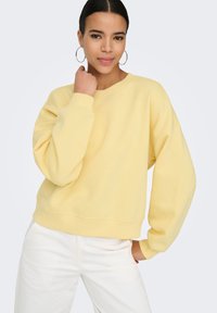 Vrouw met donker haar, gele trui en witte broek, poseert met één hand bij haar kin en de andere in haar zak, draagt oorringen.