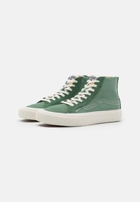 Gröna högstövlar i en slät och strukturerad materialblandning, med vita snören, en randig tygglabel och en gummisula.