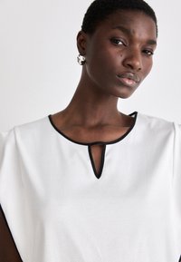 Hvit bluse med svart kragekant og en nøkkelhullsdetalj. Glatt stoff, korte ermer og et enkelt, elegang design.