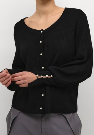 Kvinde iført en sort ribbet cardigan med perleknapper ned foran og på ærmekanterne, stylet med grå bukser.