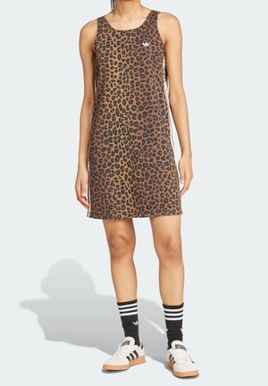 Femme portant une robe sans manches à imprimé léopard, des chaussettes Adidas noires avec des bandes blanches, et des baskets Adidas blanches avec des bandes noires.