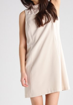 Beżowa sukienka bez rękawów z wysokim kołnierzem zdobiona różowymi koralikami, noszona przez osobę o długich brązowych włosach.