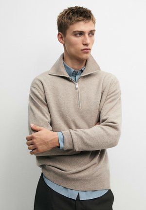 Jeune homme portant un pull beige à demi-fermeture éclair par-dessus une chemise bleue à boutons, debout les bras croisés, regardant sur le côté sur un fond uni.