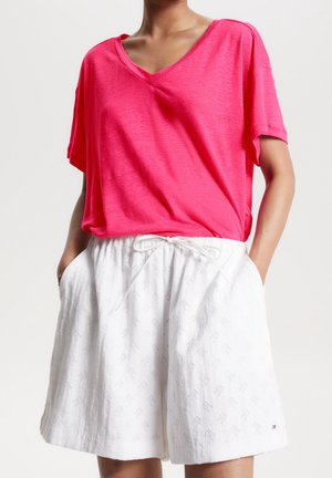 Henkilö, joka pukeutunut kirkkaan vaaleanpunaiseen V-kaula-aukkoinen lyhythihainen pusero ja valkoiset nyörit housut, joissa on hienovarainen kirjailukuvio, kädet taskuissa.