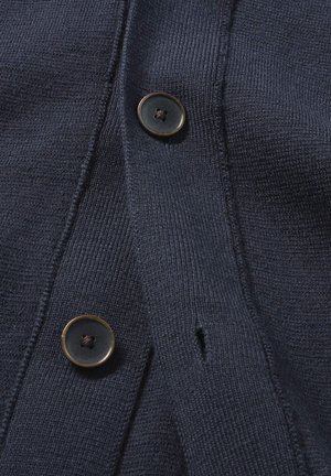 Marineblauwe gebreide cardigan met twee zwarte knopen met gouden omlijsting, gestructureerd materiaal en een klein gaatje naast de knoopsluiting.