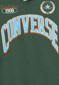 Sudadera verde con el gran texto "CONVERSE" en azul y acentos marrones. Incluye "Established 1908" y un parche de logo redondo. Textura de algodón.