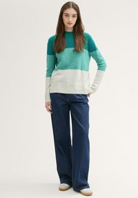 Gestrickter Pullover in Teal und Hellgrau. Lange Ärmel mit gerippten Bündchen. Eine lockere, dunkelblaue Jeans und helle Sneakers dazu.