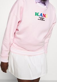 Sudadera rosa con texto multicolor "KANI" y "THE ORIGINAL" en negro debajo, combinada con una falda blanca con pliegues y puños a rayas.