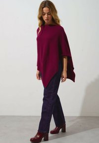 Poncho in magenta a maglia con una vestibilità comoda, orlo asimmetrico e texture a coste, abbinato a jeans dritti blu scuro e tacchi a blocco magenta.