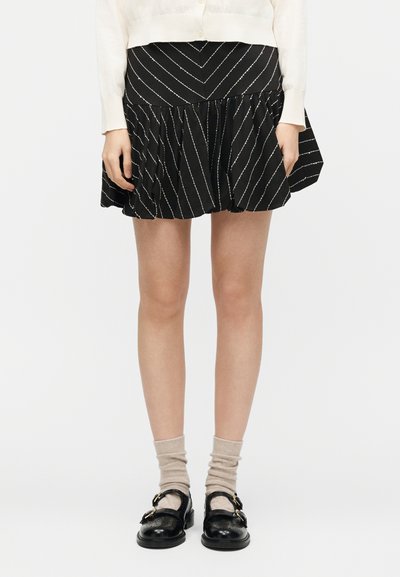 Γυναίκα με μαύρη μίνι φούστα με διαγώνιες λευκές ρίγες, μπεζ κάλτσες και μαύρα παπούτσια με αγκράφα, στέκεται μπροστά σε απλό λευκό φόντο.