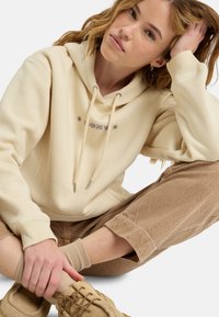 Felpa crema con cappuccio regolabile, con un logo viola e dettagli a forma di stella. Indossata con pantaloni in corduroy marroni e calzature beige.