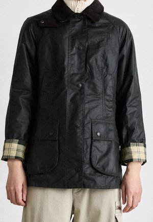 Persoon draagt een zwart gewaxed jack met een donkere kraag, knoopzakken en opgerolde mouwen die een beige geruite voering tonen, gecombineerd met een lichte broek.