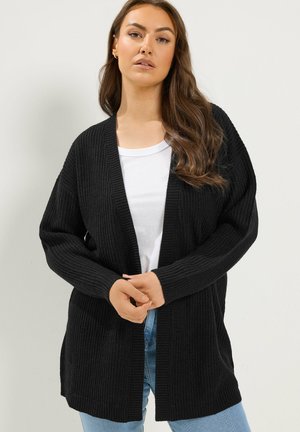 Donna con lunghi capelli castani che indossa un cardigan nero aperto sul davanti in maglia sopra una t-shirt bianca e jeans blu su sfondo semplice.