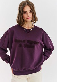 Fialová mikina vyrobená z měkké tkaniny s plyšovou, texturovanou grafikou "Jednou dávno". Má volný střih a pružné manžety.