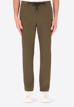 Uomo che indossa pantaloni verde oliva a gamba dritta con coulisse nera e sneakers bianche, in piedi davanti a uno sfondo semplice.