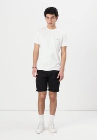 T-shirt blanche à manches courtes avec un petit logo, associée à un short noir et des baskets blanches. Les shorts ont des ourlets roulés. Design simple.