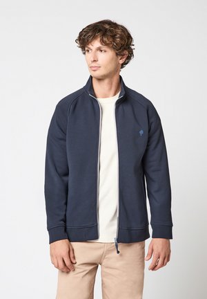 Homme portant une veste zippée bleu marine sur une chemise blanche et un pantalon beige, regardant sur le côté sur un fond uni.