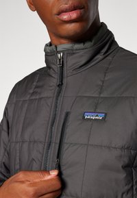 Veste matelassée noire avec un motif en losanges, fermeture éclair à l'avant, col montant et logo Patagonia blanc sur la poitrine. Texture de tissu lisse.